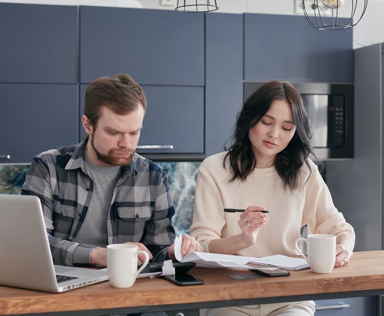A couple sits at a bright kitchen table, smiling as they review financial documents and a laptop, discussing home selling options.